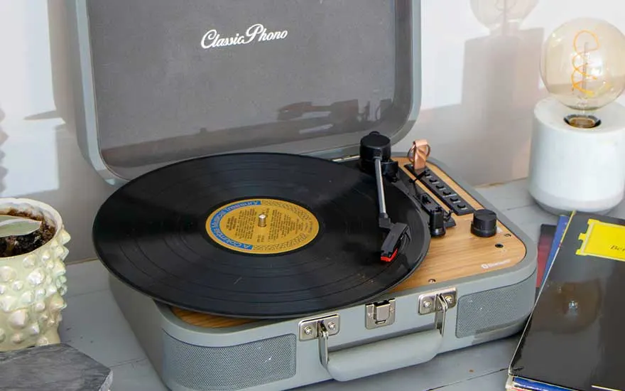 Classic Phono kofferplatenspeler in gebruik, zwarte lp draait naast een lamp en stapel platen op een stijlvol ingerichte tafel.
