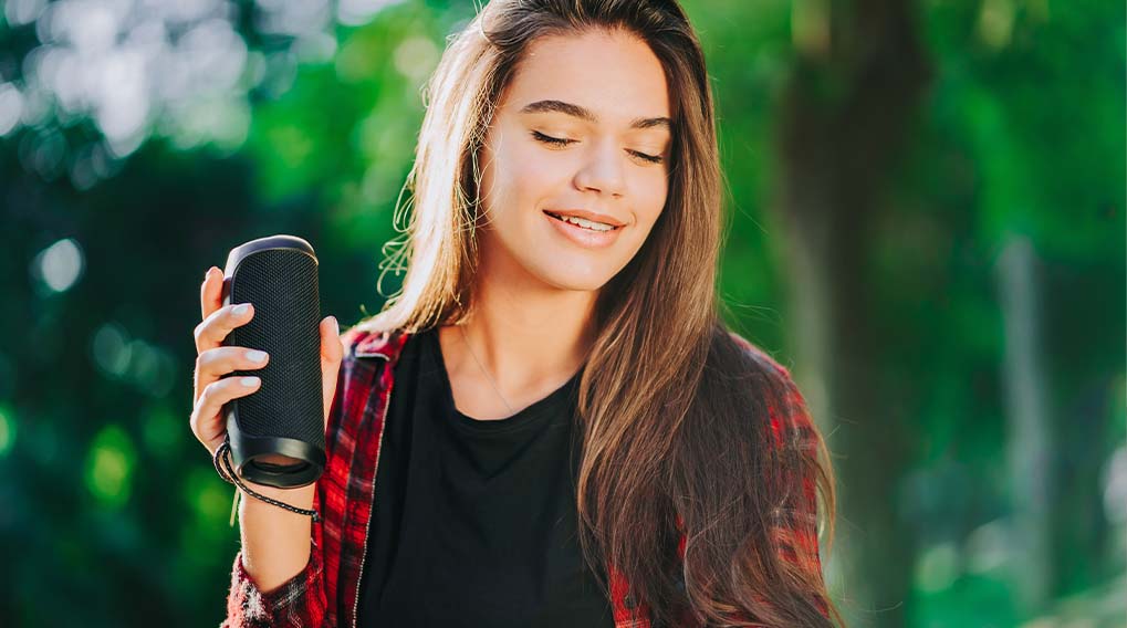 Een vrouw houdt een zwarte luidspreker vast tegen een groene achtergrond. Ze draagt een zwart shirt en een rood-zwart geruit overhemd.