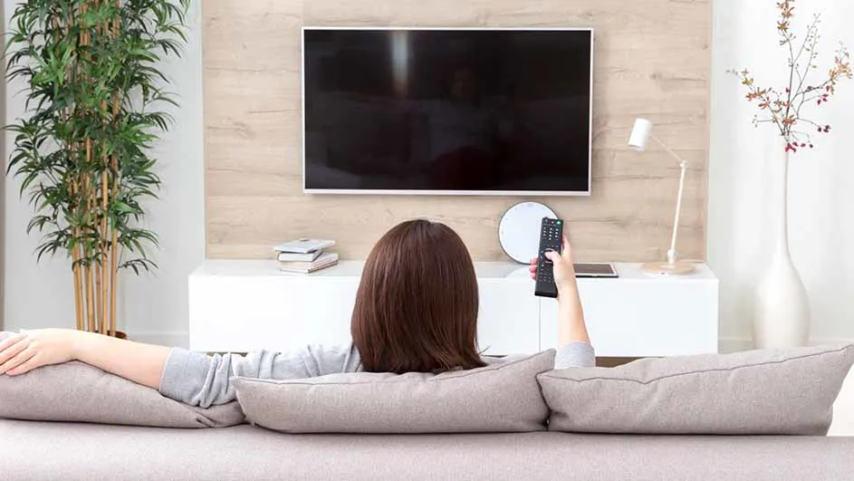 Een vrouw zit op een bank met kussens en kijkt naar een tv, met een afstandsbediening in haar hand.