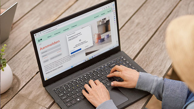 Een persoon typt op een laptop met een aanmeldingspagina op een houten tafel. Er staat een plant op de achtergrond.