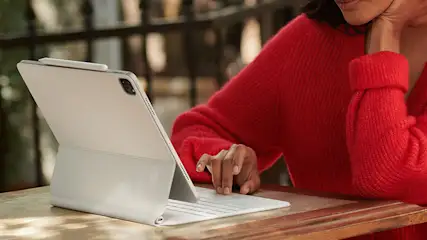 Een persoon in een rode trui gebruikt een witte tablet met een toetsenbord op een houten tafel.