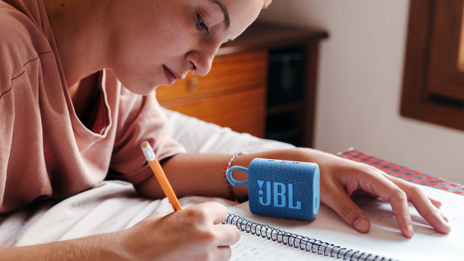 Een persoon schrijft in een notitieboek naast een blauwe JBL-luidspreker op een bed, met een houten bureau en raam op de achtergrond.