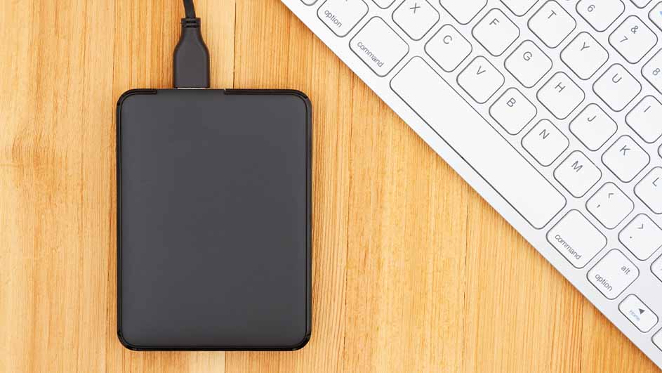 Een zwarte harde schijf met kabel ligt naast een wit toetsenbord op een houten tafel.