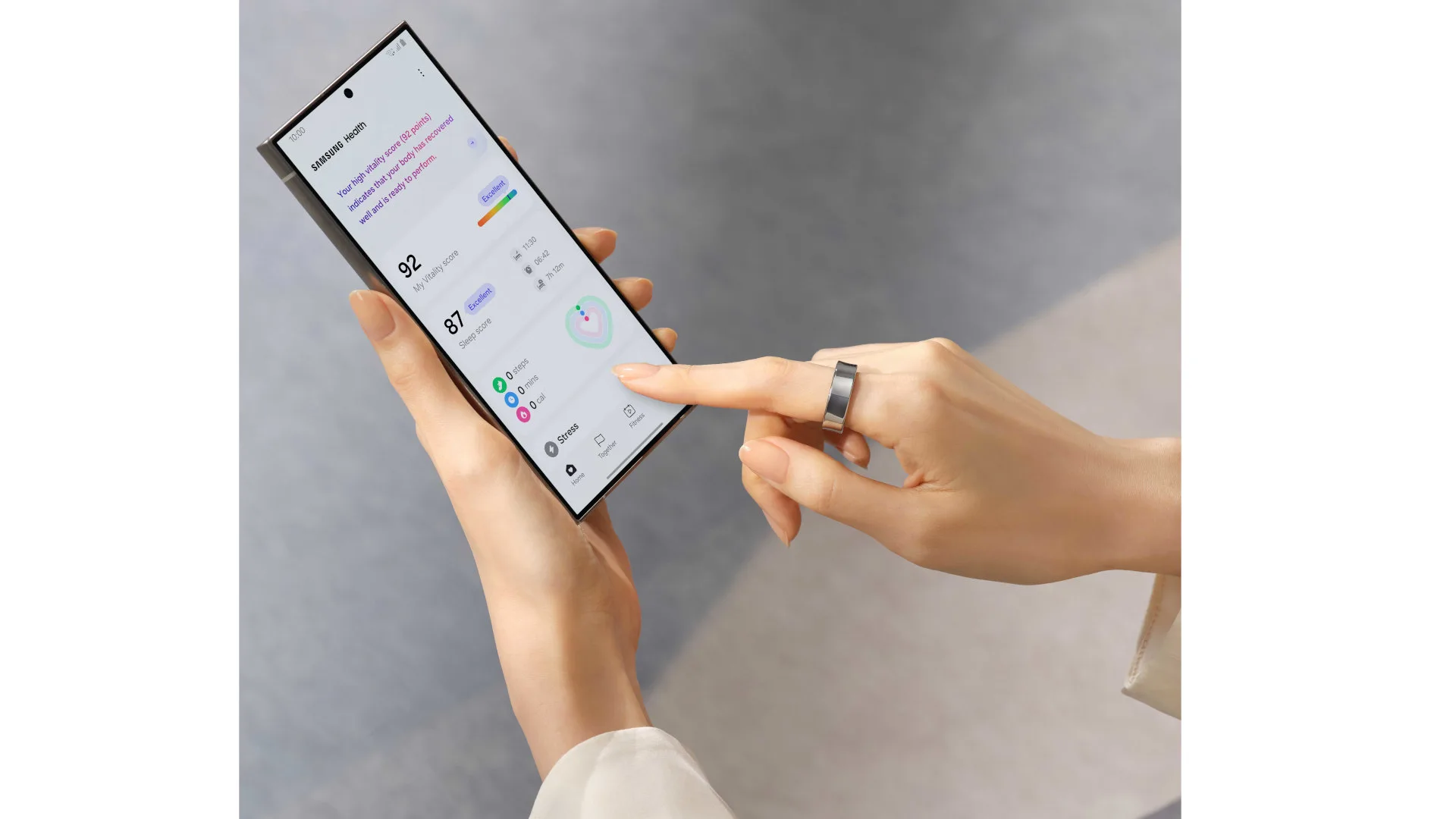 Een persoon houdt een telefoon vast met een gezondheidsapp en draagt een zilveren ring. De achtergrond is grijs.