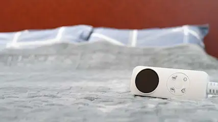 Een wit bed met blauwe kussens en een rode achterwand. Een witte bedieningseenheid met een ronde, zwarte knop ligt op het bed.