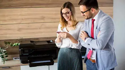 Twee mensen kijken naar een telefoon bij een printer. De man draagt een pak en de vrouw een plooirok.