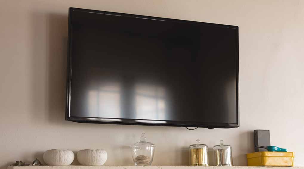 Black flat-screen TV is mounted on a beige wall above a shelf with decorative white vases and glass jars.