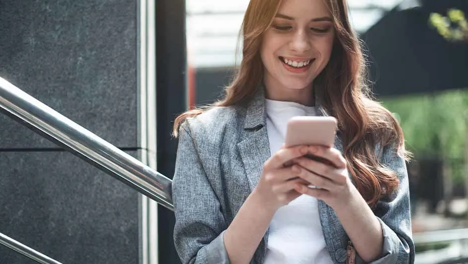 Vrouw glimlacht en kijkt naar roze smartphone. Ze draagt een grijs jasje en een wit shirt, staat bij een metalen reling.