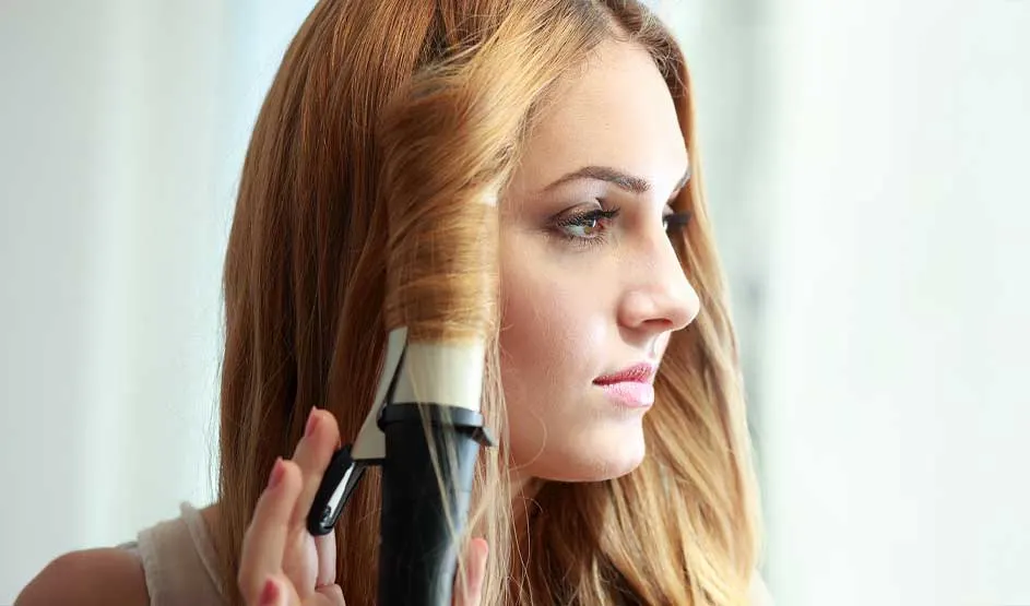 A young woman curls a blonde strand with a curling iron; she gazes right in soft daylight, with a blurred background.