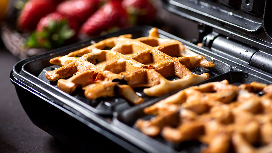 Twee goudbruine wafels in een zwart wafelijzer, met aardbeien op de achtergrond.
