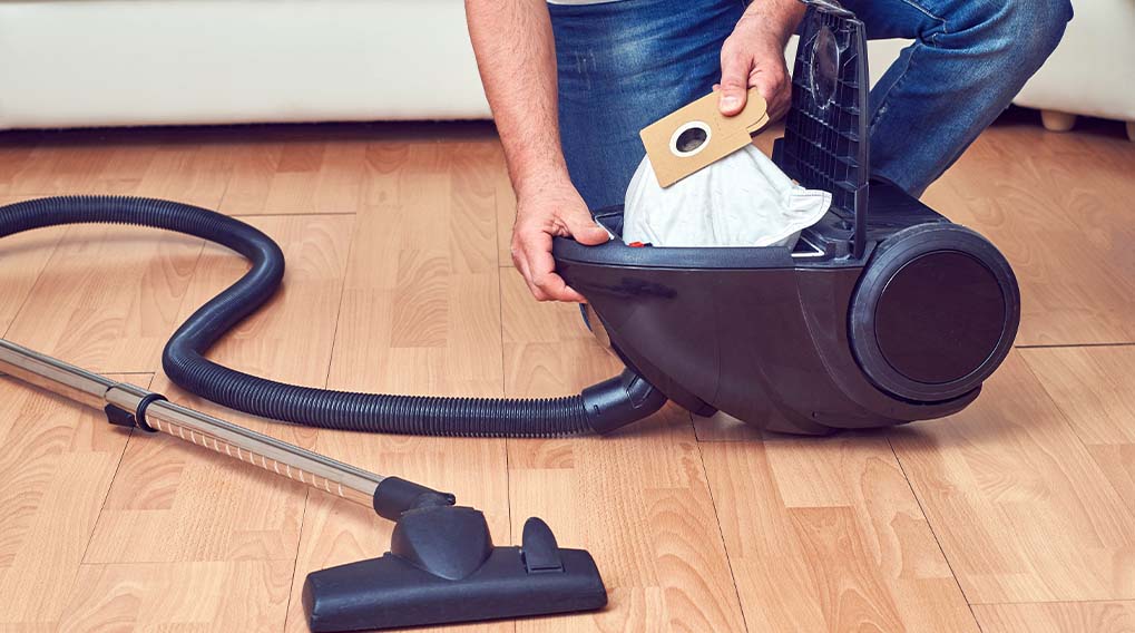 Een persoon vervangt een stofzuigerzak in een zwarte stofzuiger op een houten vloer, met een slang en zuigmond zichtbaar.