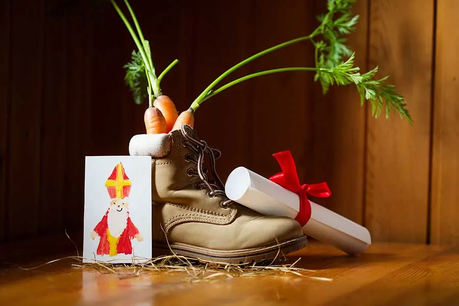 Een schoen met wortels erin, een tekening van Sinterklaas en een opgerolde brief met een rood lint, op een houten vloer.