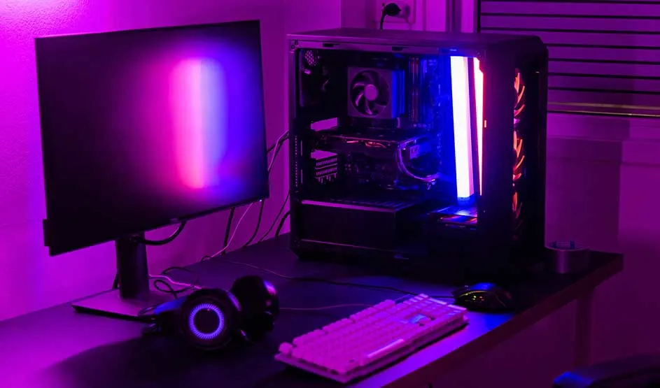 Gaming setup with monitor, PC tower glowing in purple neon, white keyboard, mouse and headphones on a sleek dark desk surface.