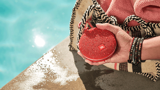 Een hand houdt een rode JBL-luidspreker vast naast een zwembad, met een tas en handdoek op de achtergrond.