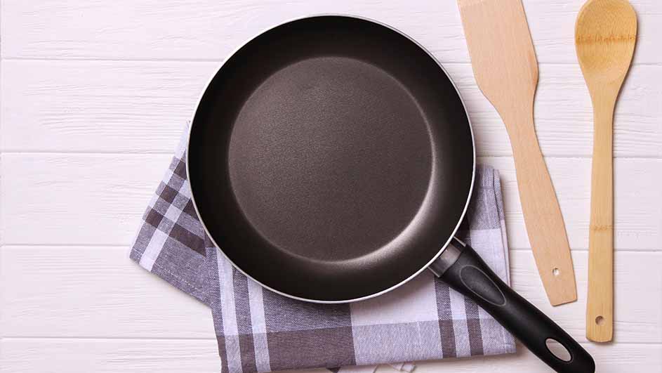 Een zwarte koekenpan op een geruite doek, naast houten spatels op een witte houten achtergrond.