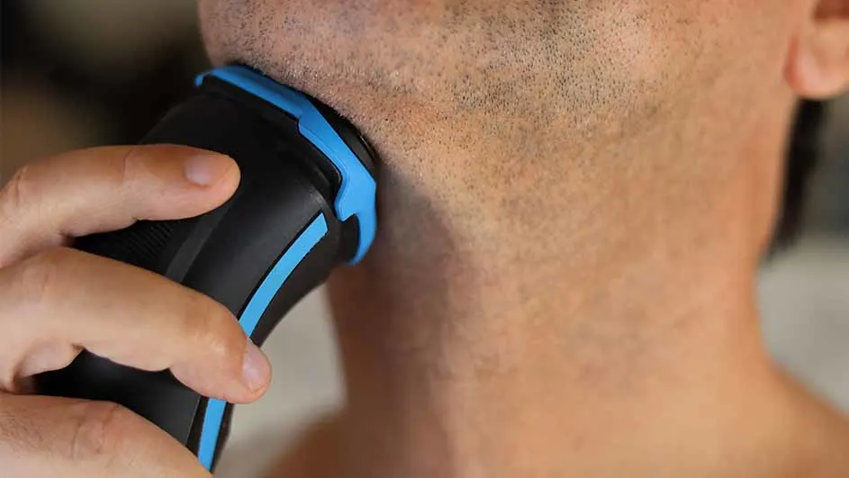 Een man scheert zijn gezicht met een zwart en blauw elektrisch scheerapparaat.