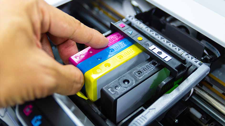 Een hand plaatst roze, blauwe en gele inktpatronen in een printer met zwarte en grijze onderdelen.