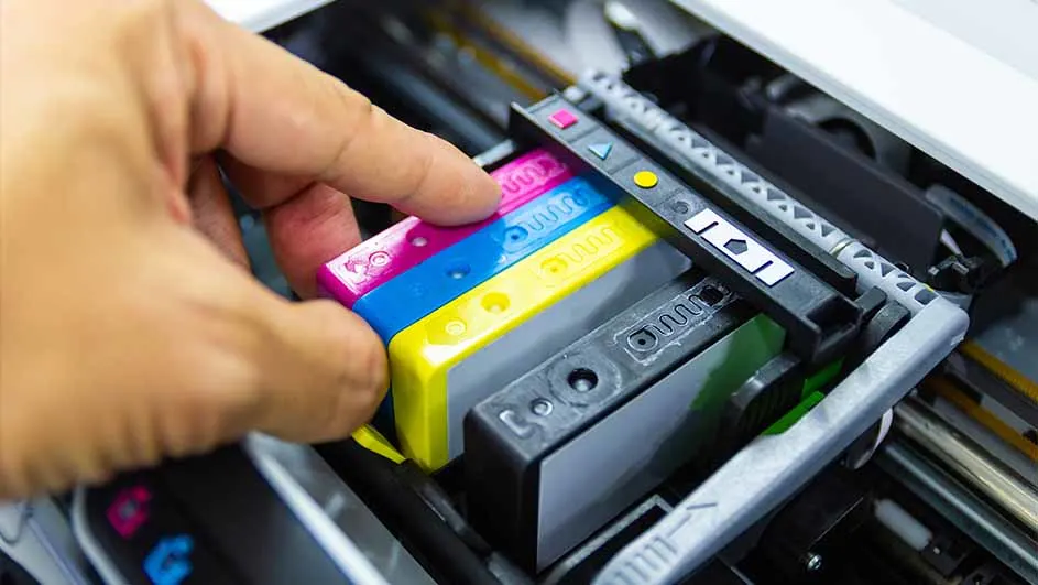 Een hand plaatst roze, blauwe en gele inktpatronen in een printer met zwarte en grijze onderdelen.