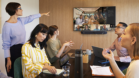 Groep medewerkers rond vergadertafel volgt hybride meeting op groot scherm, terwijl een collega vooraan iets toelicht.