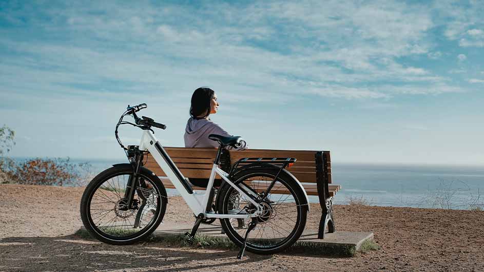 Vrouw zit op een bank bij een witte elektrische fiets, met uitzicht op de blauwe zee en de hemel.
