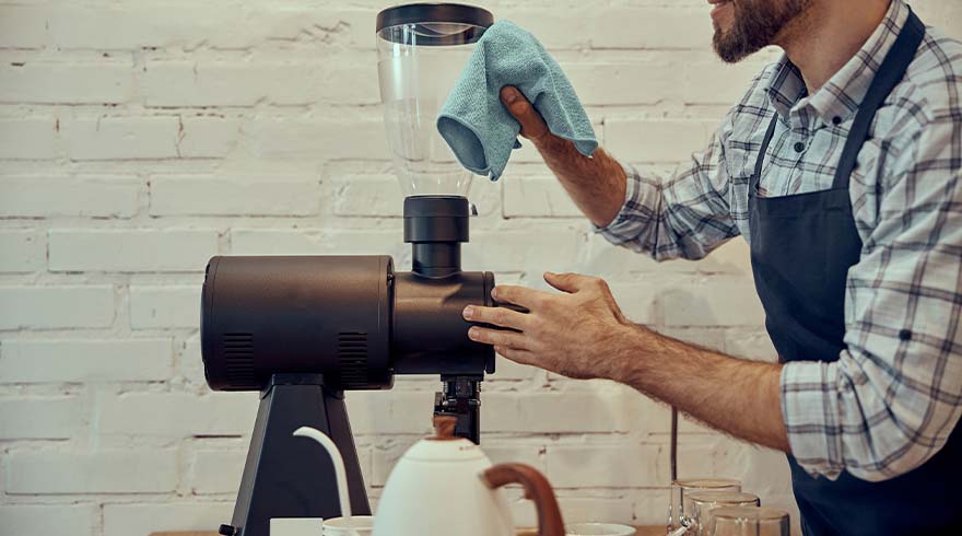 Een persoon in een schort maakt een zwarte koffiemolen schoon met een blauwe doek tegen een witte bakstenen muur.