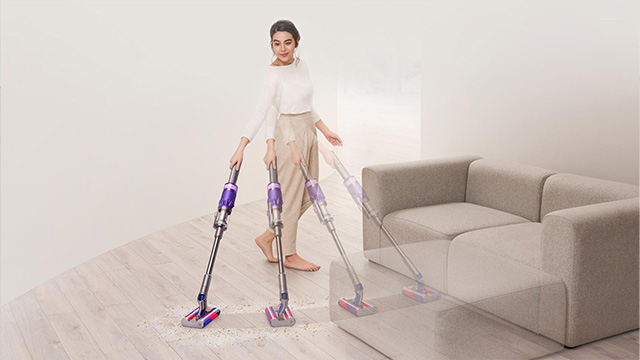 Een vrouw stofzuigt een houten vloer met een paarse steelstofzuiger, naast een beige bank in een lichte ruimte.