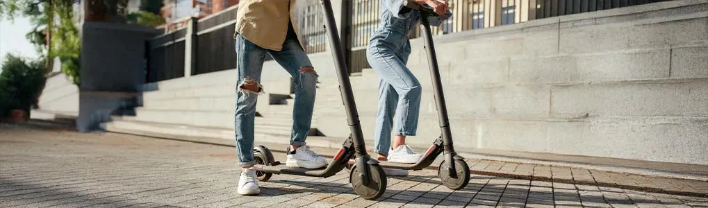 Twee personen op elektrische steps, vastgelegd vanaf kniehoogte. Ze dragen casual kleding met sneakers en staan op een bestrate straat met een stenen trap en hekwerk op de achtergrond. De sfeer oogt stads en modern.