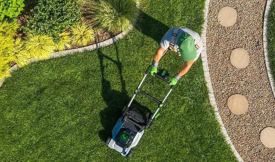 Een persoon met groene pet maait het gras met een elektrische grasmaaier naast een pad met ronde tegels. Geen merk zichtbaar.