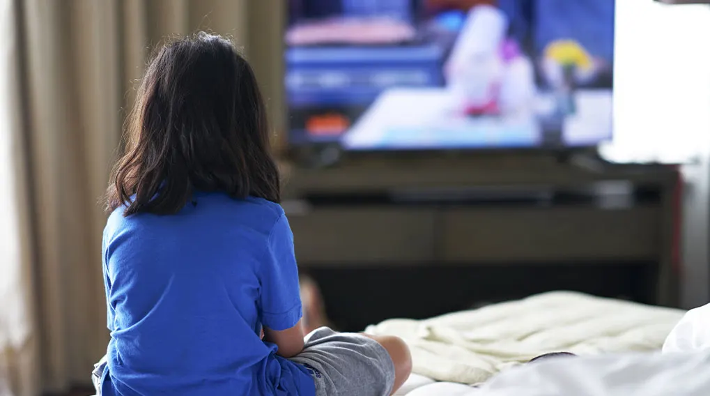 Een kind zit op een bed met een blauw shirt en grijze korte broek, kijkend naar een tv in een kamer.