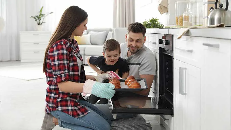 Een gezin kijkt naar broodjes in een oven. De vrouw draagt ovenwanten en een rood geruit overhemd. Keuken op de achtergrond.