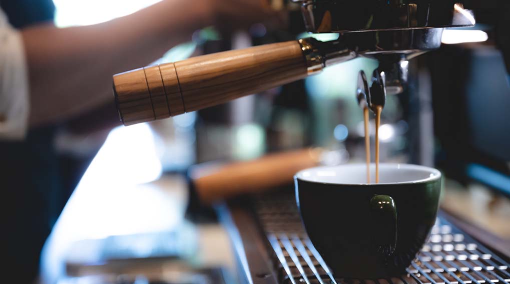 Een espressoapparaat vult een donkergroene kop met espresso op een metalen rooster. Een hand is zichtbaar op de achtergrond.