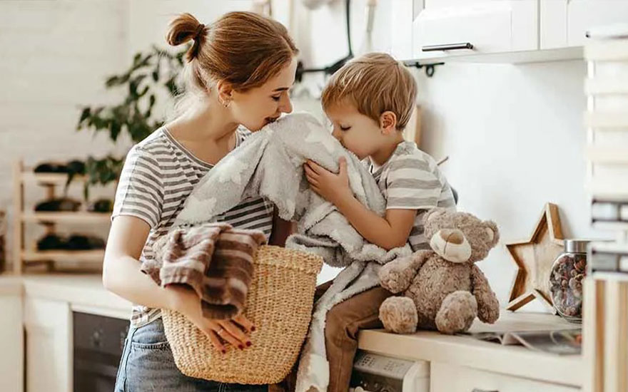 A mother and child are smelling a blanket in a bright kitchen. The mother's wearing a striped top and jeans, while the child is sitting on the worktop with a teddy bear. A laundry basket is next to them