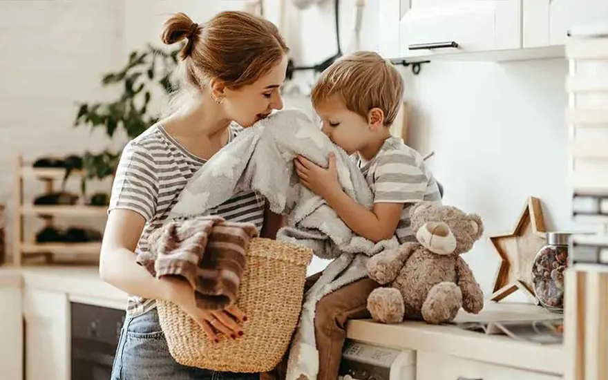 A mother and child are smelling a blanket in a bright kitchen. The mother's wearing a striped top and jeans, while the child is sitting on the worktop with a teddy bear. A laundry basket is next to them