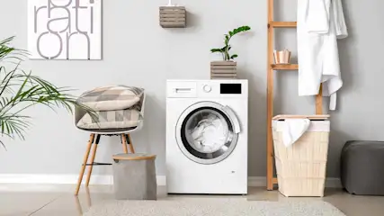 Een wasruimte met een witte wasmachine, een houten ladder met witte handdoeken en een stoel met kussens.