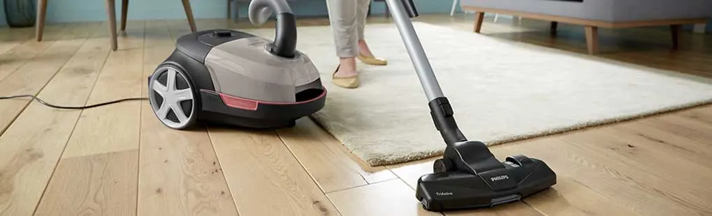 Someone is vacuuming the light floor in a living room with a cylinder vacuum cleaner; a yellow sofa and white table are in the background.