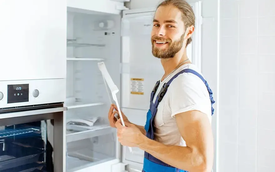 Monteur in blauwe overall lacht naar de camera terwijl hij een open inbouwkoelkast inspecteert en een wit onderdeel of plank in zijn handen houdt