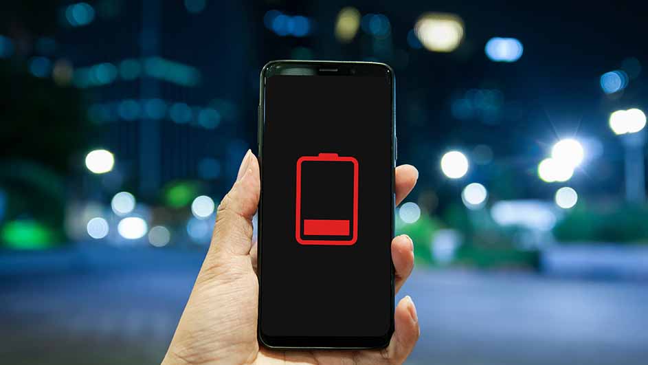 Een hand houdt een telefoon vast met een rood batterijsymbool op een zwart scherm, tegen een vage stadsachtergrond.