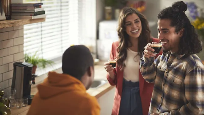 Drie mensen in een keuken drinken koffie, lachend en genietend van het moment. Een koffiezetapparaat staat op het aanrecht.