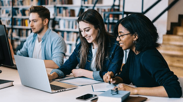 Drie studenten werken aan een tafel met een laptop en computer in een bibliotheek met boeken op de achtergrond.