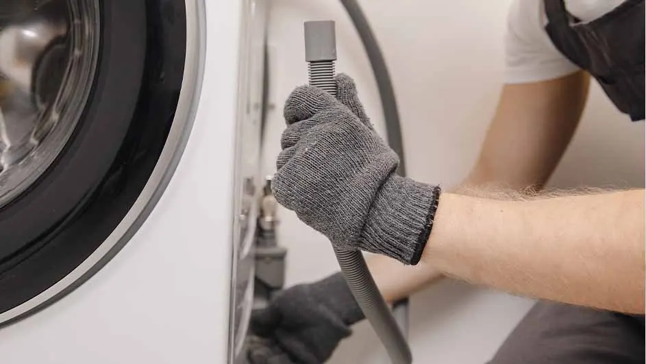 A gloved hand holds a gray hose connected to a white washing machine, with part of a person visible on the right.