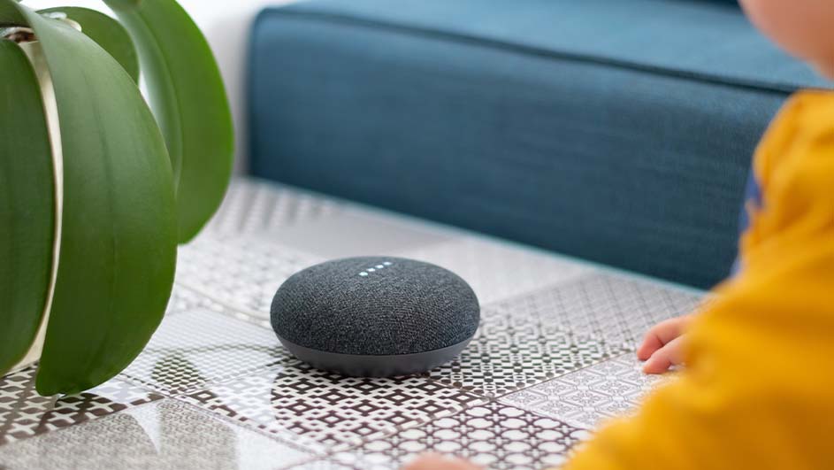 Grijze slimme speaker op een tegelvloer, groene plant links, blauwe bank op de achtergrond en de hand van een kind rechts.