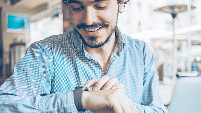 Een man met een blauw overhemd kijkt naar een smartwatch. De achtergrond toont een wazig interieur met een lamp en een kunstwerk.