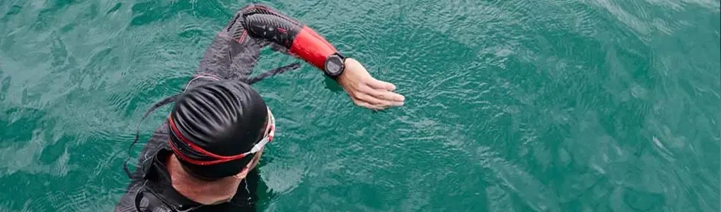 A swimmer in a black cap and wetsuit swims in blue-green water, arm extended. The swimmer wears goggles and a watch.