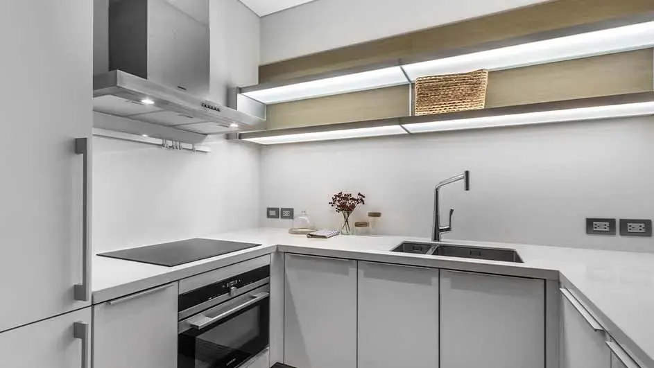 Strakke hoekkeuken met witte kasten, inductiekookplaat, rvs‑afzuigkap, spoelbak en lange verlichte legplanken boven het blad.