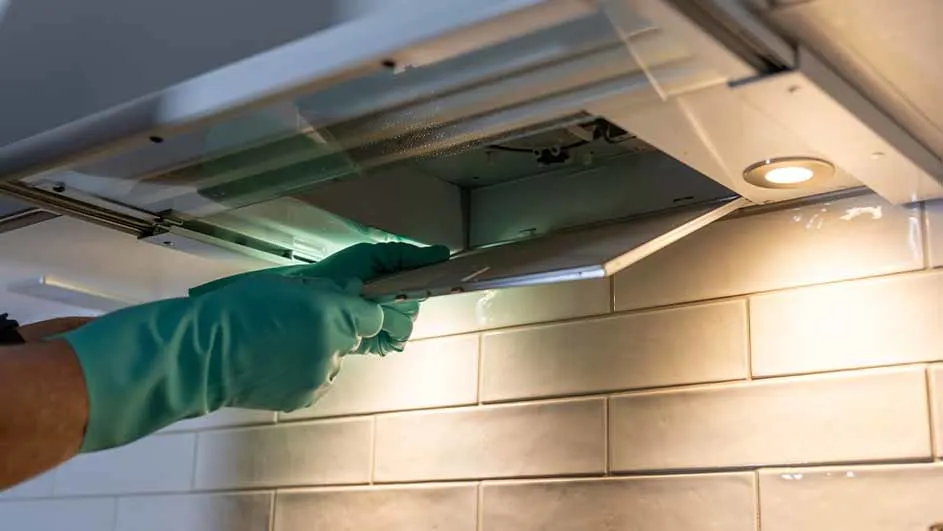 Iemand met groene handschoenen verwijdert een filter uit een witte afzuigkap boven een witte tegelwand.