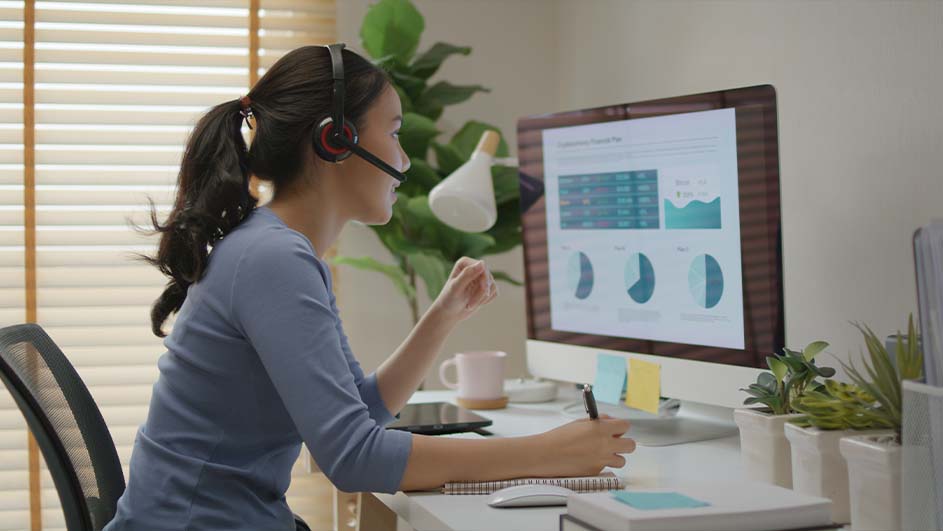 Vrouw met headset kijkt naar een computerscherm met grafieken, schrijft in een notitieboek op een wit bureau. Planten op achtergrond.