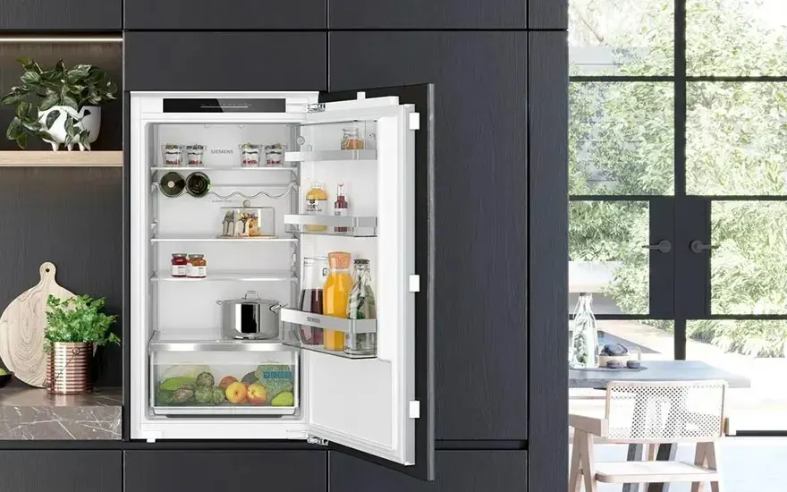 Een ingebouwde Siemens koelkast staat open in een moderne keuken, gevuld met groenten, fruit, flessen, potjes en een pan. Alles netjes ingedeeld.