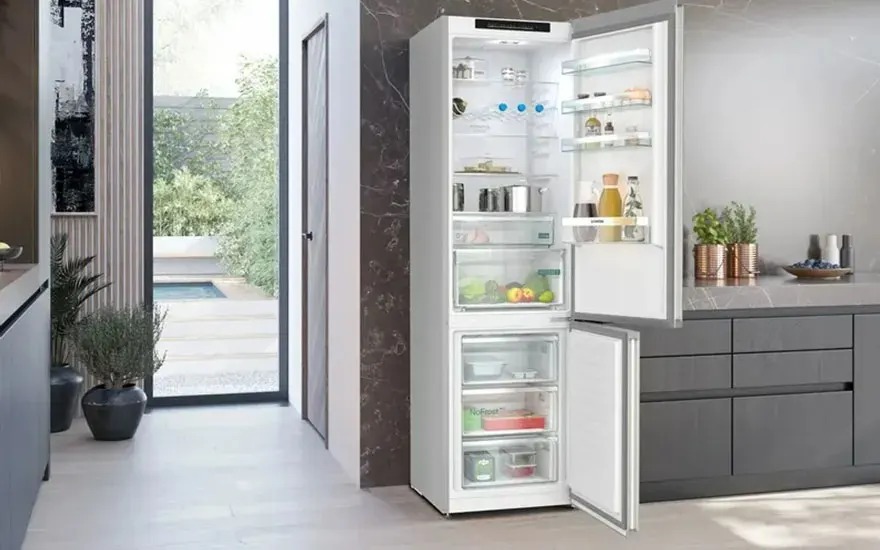 In a modern kitchen a tall fridge freezer stands open, neatly organised with food, drinks and fresh vegetables on clear shelves.