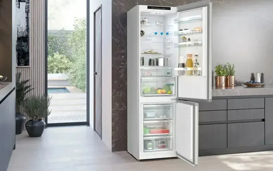 In a modern kitchen a tall fridge freezer stands open, neatly organised with food, drinks and fresh vegetables on clear shelves.