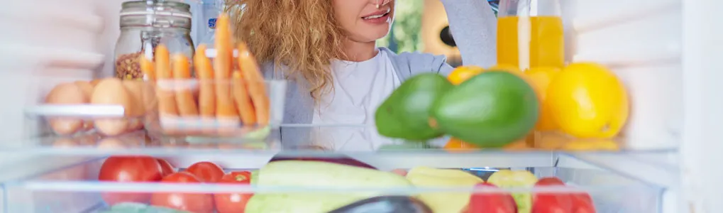 Vrouw fronst haar neus voor een open koelkast vol groenten en sap, waarschijnlijk vanwege een onaangename geur uit de koelkast.